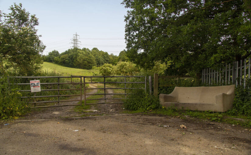 A fly-tipped sofa in the countryside next to a fence that bears a sign warning against fly-tipping