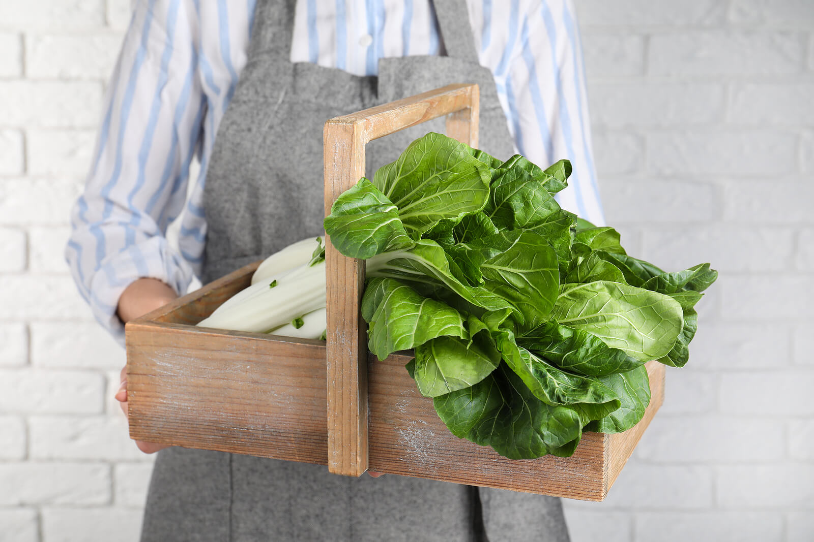 A reusable wooden food crate full of vegetables, being held by a woman wearing an apron.