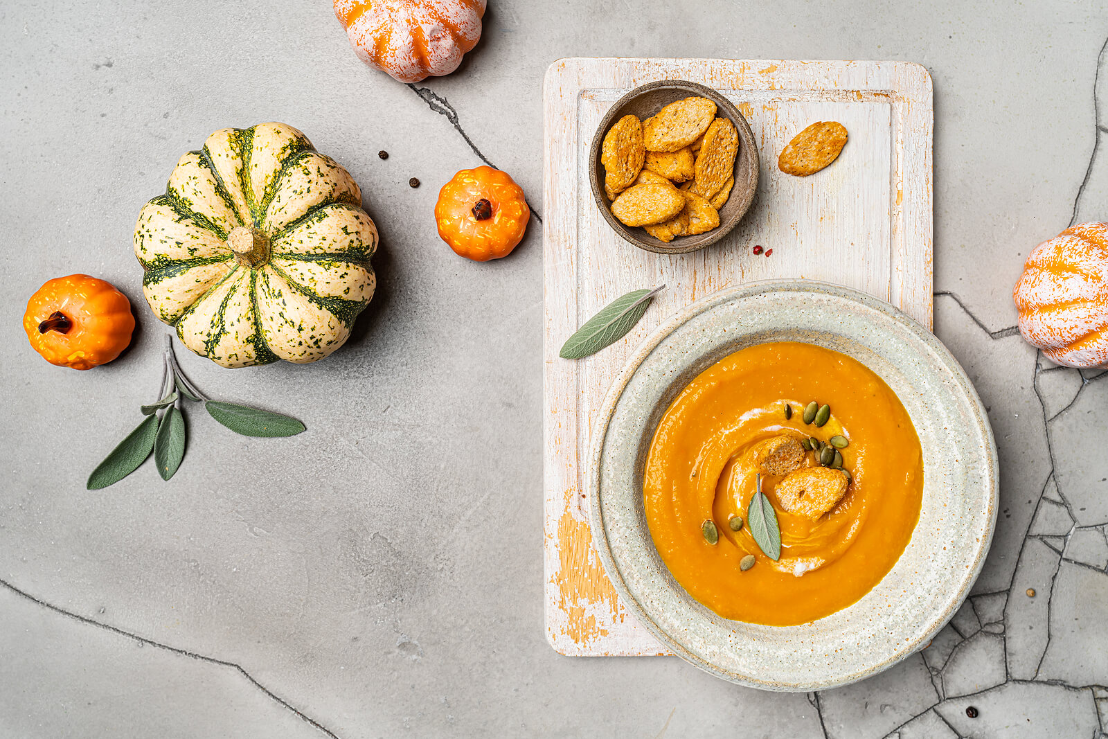 A flat lay featuring pumpkins and squash, and a bowl of pumpkin soup sat on top of a chopping board.
