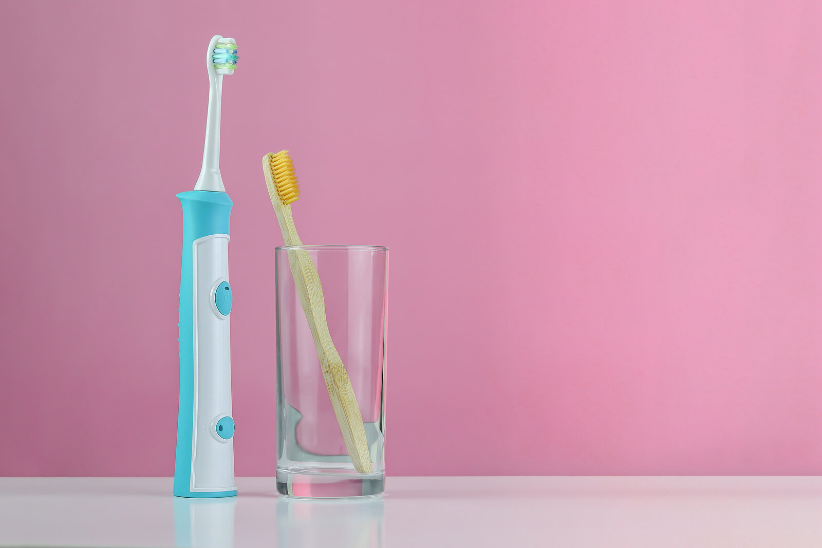 An electric toothbrush sat on a surface next to a glass containing a bamboo toothbrush.